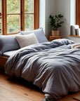 Neatly made bed with gray bedding in a bright bedroom with large windows.