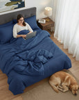 Woman using a laptop on a bed with navy blue cotton sateen bedding, dog lying on the floor beside her.