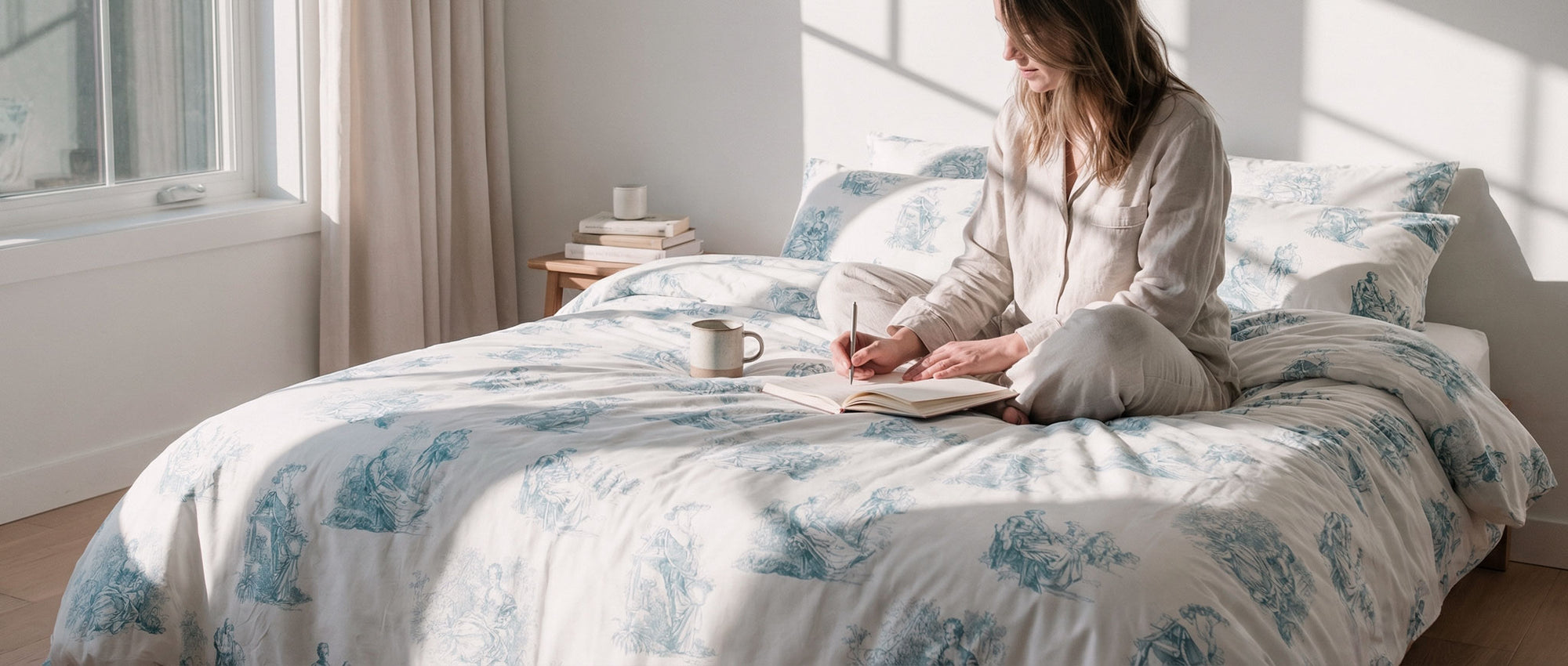 A woman sitting and writing on a bed covered with a blue floral toile de jouy duvet cover bedding set