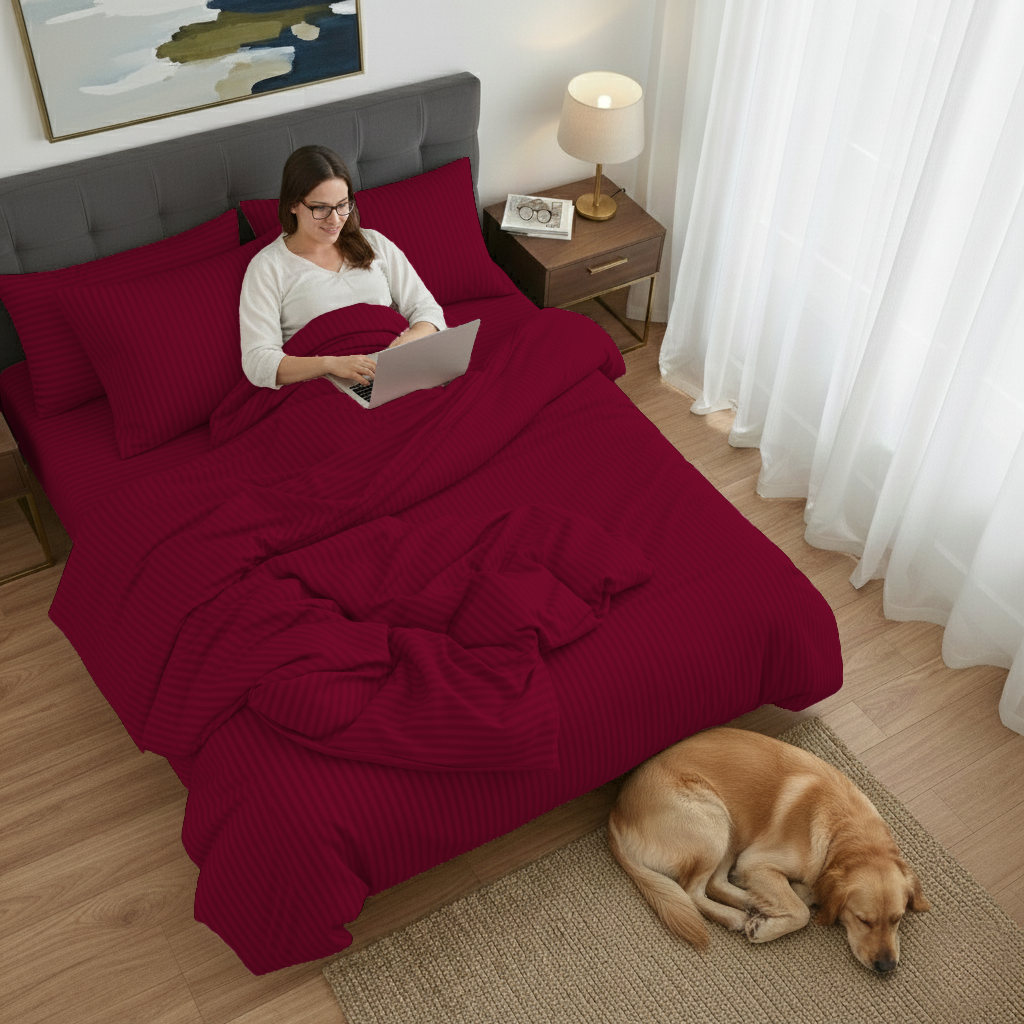 Woman using a laptop on a bed with red bedding, dog sleeping on the floor.