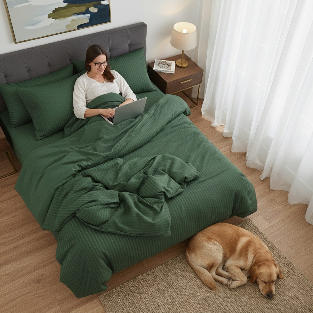 Woman using a laptop on a bed with green cotton sateen bedding, dog lying on the floor beside her.