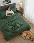 Woman using a laptop on a bed with green cotton sateen bedding, dog lying on the floor beside her.