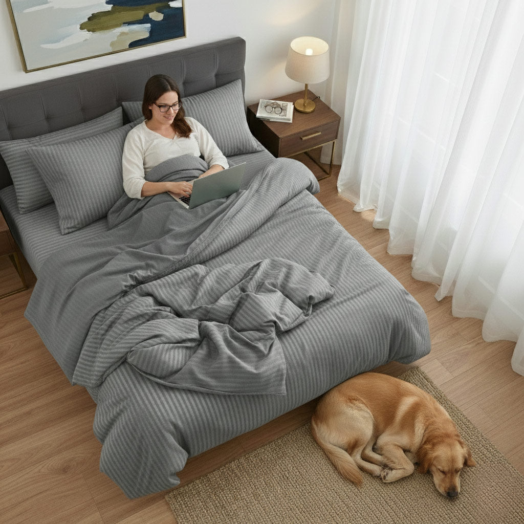 Woman using a laptop on a bed with grey cotton sateen bedding, dog lying on the floor beside her.