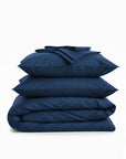 Stack of navy blue cotton sateen bedding bundle including a duvet cover, two pillowcases, and a flat sheet on a white background