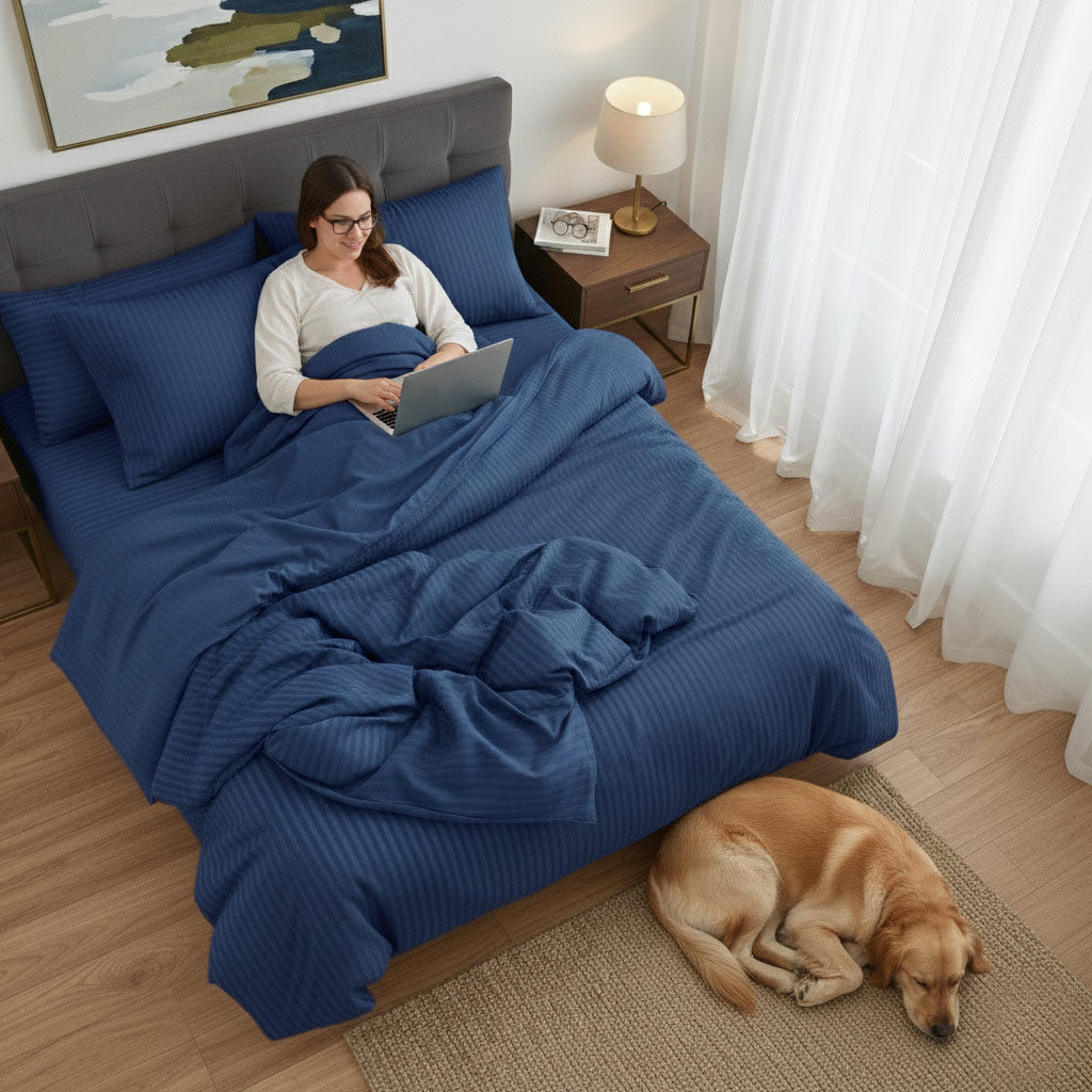 Woman using a laptop on a bed with navy blue cotton sateen bedding, dog lying on the floor beside her.