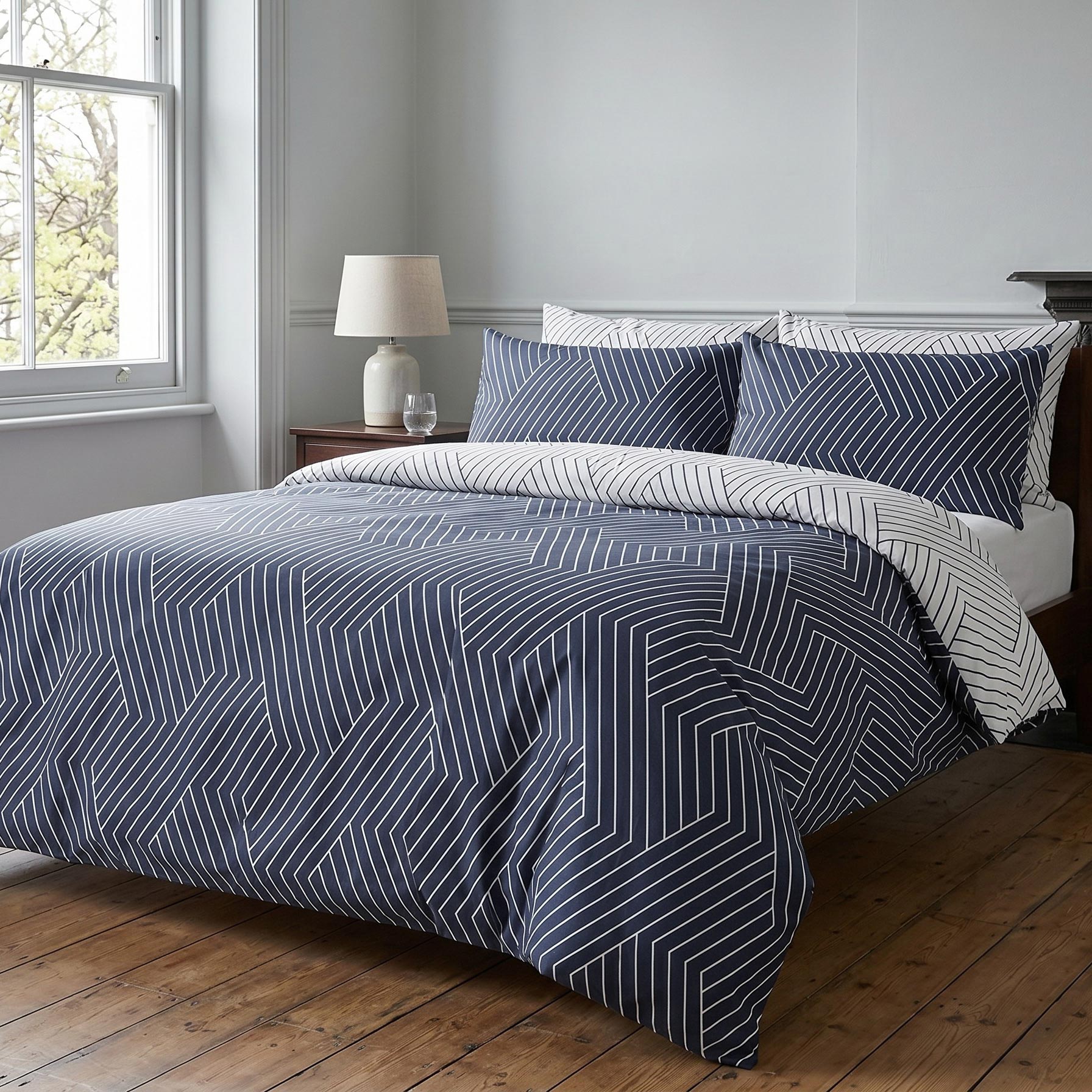 A navy blue and white geometric patterned bedding set, including a duvet cover and matching pillowcases, styled on a bed in a bedroom with a wooden floor, bedside table, and window.
